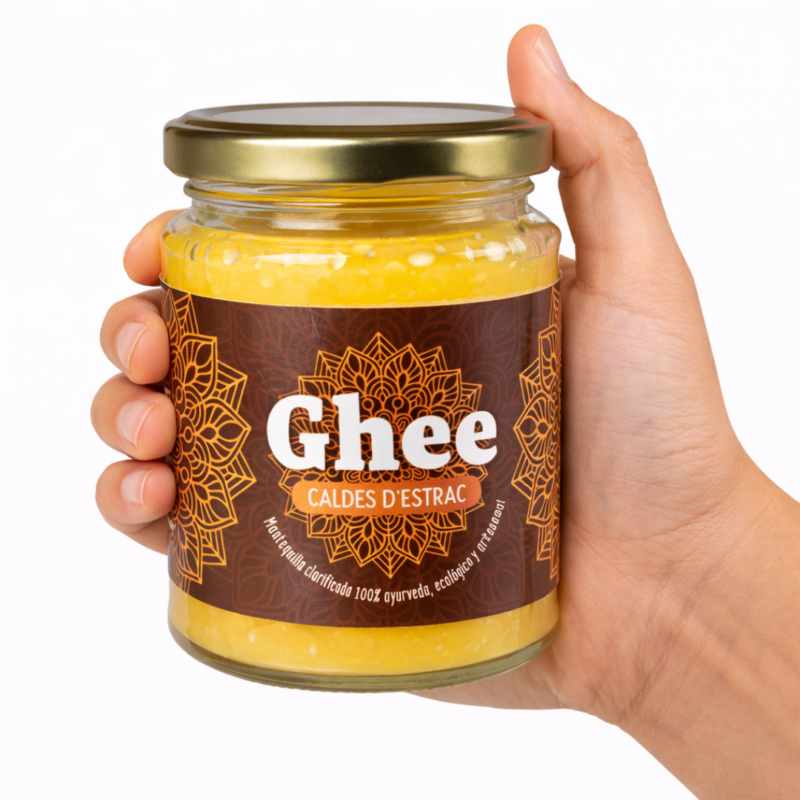 Mano sosteniendo un tarro grande de ghee Caldes d’Estrac con etiqueta marrón y diseño mandala sobre fondo blanco.