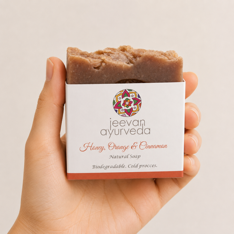 Mano neutra sosteniendo jabón natural Jeevan Ayurveda Honey, Orange & Cinnamon sobre fondo blanco minimalista.