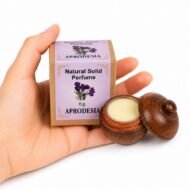 Mano sosteniendo el Perfume Sólido Aprodesia Jains de 6 g con envase de madera y caja original sobre fondo blanco.