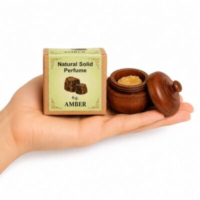 Mano sosteniendo el Perfume Sólido Amber de Jains de 6 g con envase de madera y caja original sobre fondo blanco.